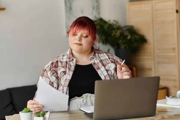 A woman in a plaid shirt sits at a desk, reviewing documents and using a laptop. — Stock Photo