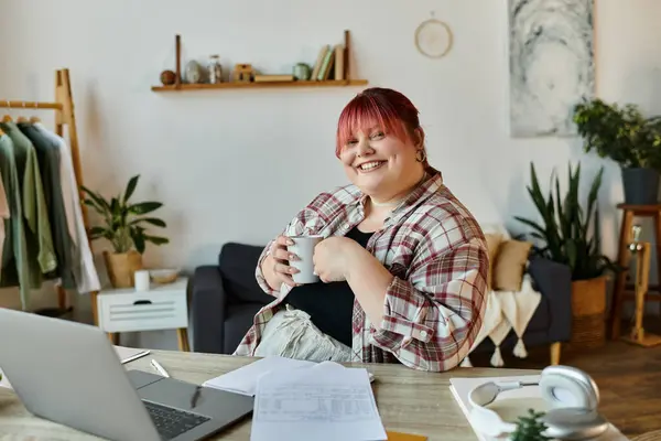 A woman sits at a desk with a mug in her hand, looking towards the camera and smiling. — Stock Photo