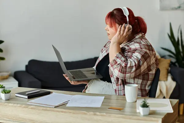 A woman wearing headphones sits at a desk, holding a laptop while looking at the screen. — Stock Photo