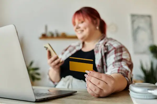 A woman holds a credit card and a phone while smiling at a laptop. — Stock Photo