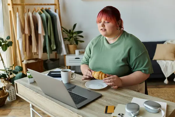Una mujer de talla grande se toma un descanso de su trabajo en casa, disfrutando de una pastelería y una taza de café. - foto de stock