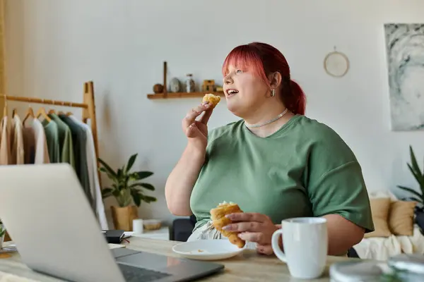 A woman with red hair eats a pastry while sitting at her kitchen table. — Stock Photo