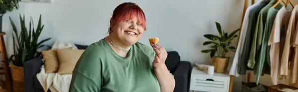 Una mujer de talla grande con cabello rosa disfruta de un dulce capricho en su acogedor salón. - foto de stock