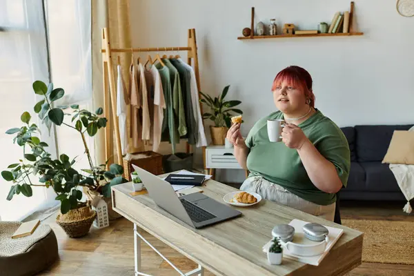 A woman enjoys a coffee and pastry while working at her desk. — Stock Photo