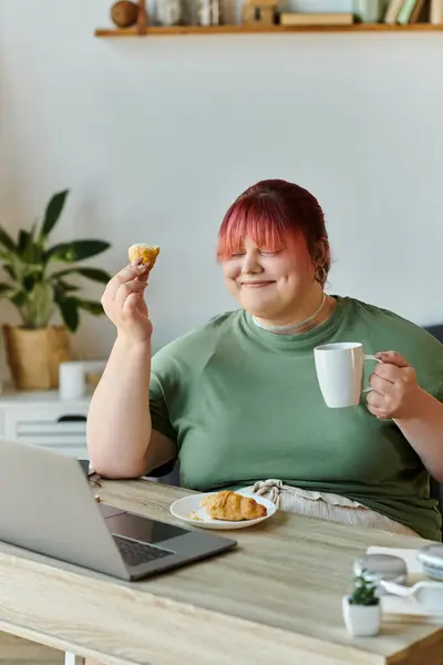 Eine Frau mit rosa Haaren lächelt, während sie zu Hause ein Gebäck und eine Tasse Kaffee genießt. — Stockfoto