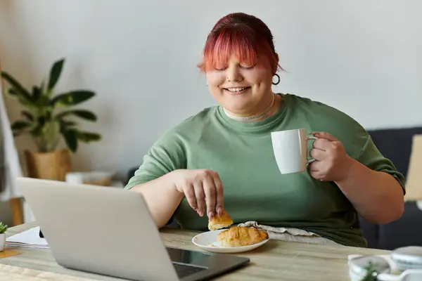 Una mujer de talla grande sonríe mientras disfruta del café y la pastelería mientras trabaja en una computadora portátil. — Stock Photo