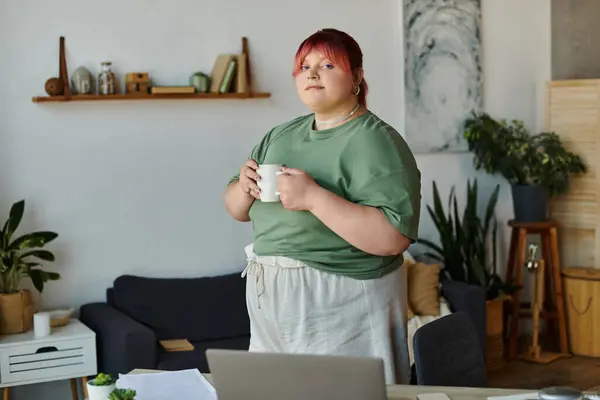 A woman stands in her home office, holding a mug and looking thoughtfully. — Stock Photo