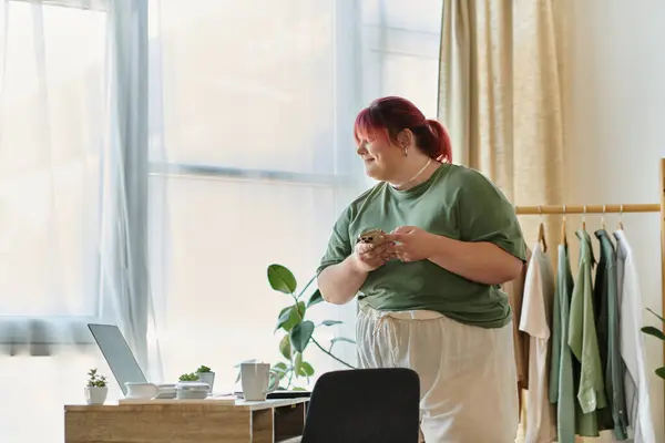 Женщина plus-size с рыжими волосами улыбается, глядя в окно, держа в руках телефон. — стоковое фото