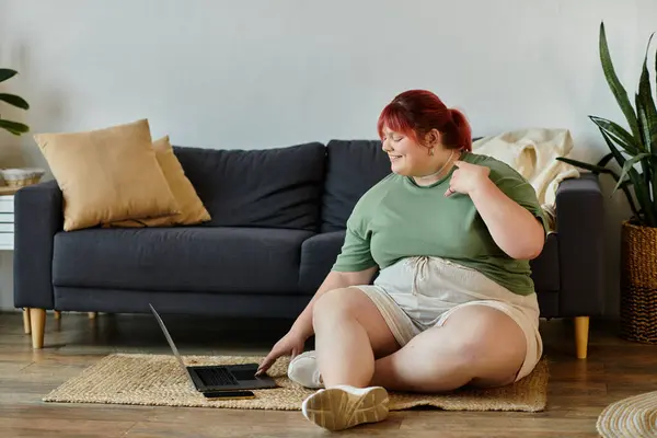 A plus size woman with red hair sits on a rug, smiling and using a laptop. — Stock Photo
