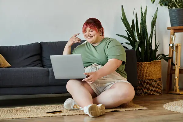 Eine Frau mit rosa Haaren sitzt auf dem Boden vor einem Sofa und lächelt, während sie an ihrem Laptop arbeitet.. — Stockfoto