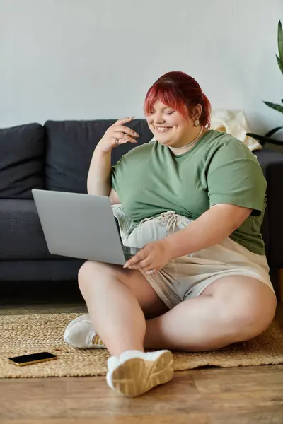 A woman with red hair sits on the floor using a laptop while smiling. — Stock Photo