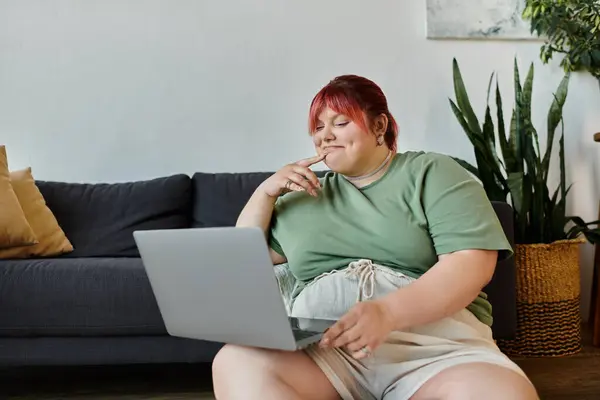 Eine Plus-Size-Frau sitzt auf einer Couch und blickt nachdenklich auf einen Laptop. — Stockfoto