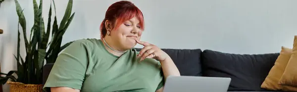 Женщина plus size с розовыми волосами сидит на диване в своем доме, улыбаясь, глядя на ноутбук. — стоковое фото