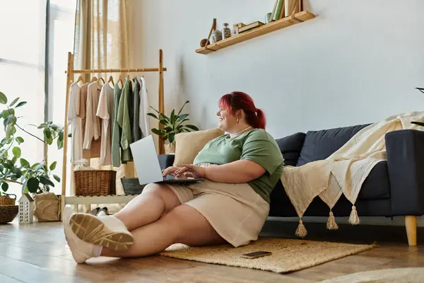 Donna in camicia verde e pantaloncini bianchi si siede sul pavimento, utilizzando il computer portatile di fronte al divano — Foto stock