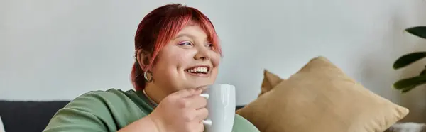 A woman with pink hair smiles and enjoys a drink while sitting on a couch. — Stock Photo