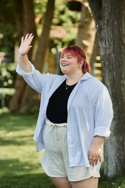 A woman with red hair waves to someone off-camera, smiling brightly in a green park. — Stock Photo