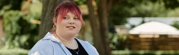 Una mujer con el pelo rosa sonríe y mira a la cámara en un entorno soleado al aire libre. - foto de stock