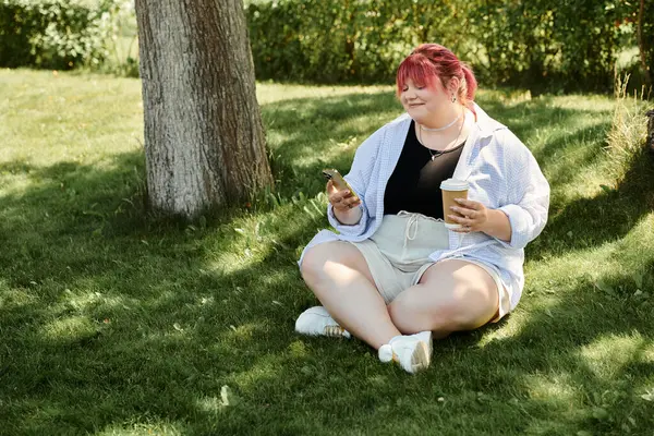 A woman sits on the grass, enjoying a coffee and browsing her phone. — Stock Photo