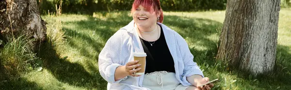 Una mujer se sienta junto a un árbol, sonriendo mientras sostiene una taza de café. - foto de stock