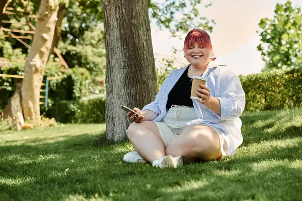 A woman sits on the grass in a park, smiling and holding a cup of coffee. — Stock Photo