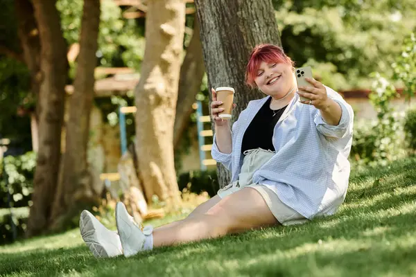 Una donna si siede in un parco, sorridendo e scattando un selfie con il telefono mentre tiene in mano una tazza di caffè. — Foto stock
