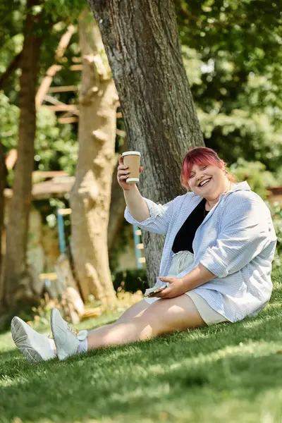 Una mujer con el pelo rosa se sienta en la hierba, sonriendo y sosteniendo una taza de café. - foto de stock