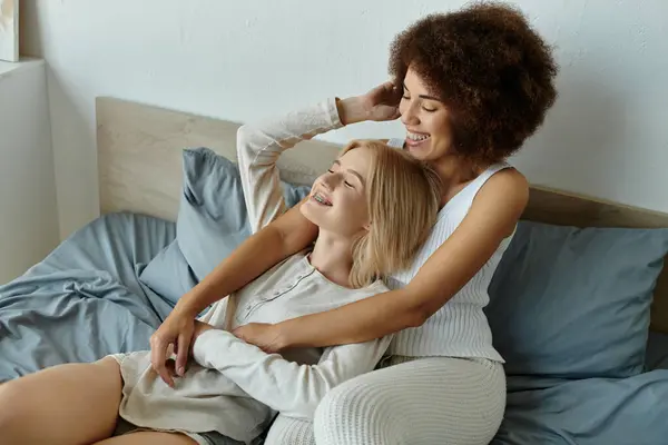 Ein multikulturelles lesbisches Paar teilt eine herzliche Umarmung auf seinem Bett, gekleidet in kuschelige Homewear und strahlende Freude. — Stockfoto