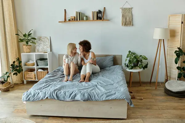 Two women in cozy homewear sit on a bed, facing each other and enjoying a moment together. — Stock Photo