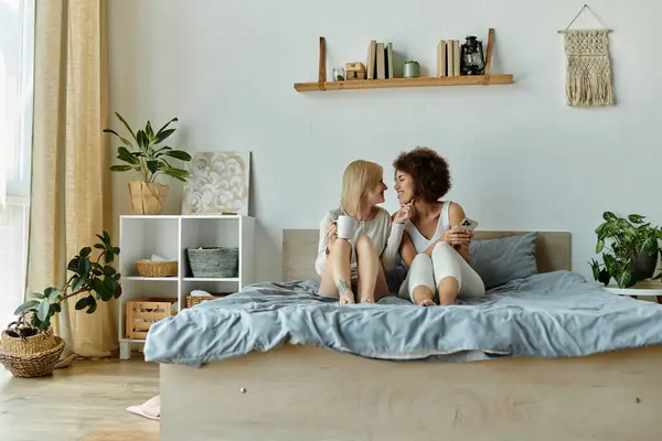 Две женщины, одетые в уютную домашнюю одежду, разделяют утренний момент вместе, сидя на кровати и улыбаясь друг другу. — стоковое фото