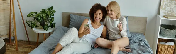 Ein multikulturelles lesbisches Paar kuschelt im Bett, eine blickt auf ihr Handy, während die andere an einem Becher nippt, beide lachen. — Stockfoto