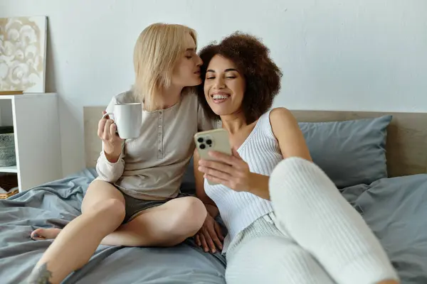 A lesbian couple shares a tender kiss and a cup of coffee while scrolling through their phone on a cozy morning. — Stock Photo