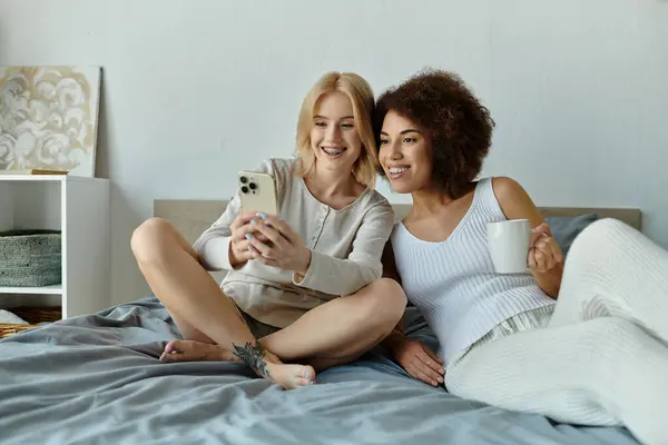 A lesbian couple cuddles in bed, looking at a phone and enjoying a cup of coffee. — Stock Photo