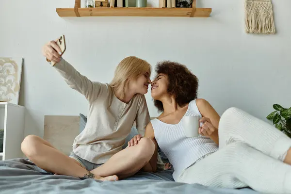 A multicultural lesbian couple enjoys a cozy moment at home, taking a selfie while sharing a warm drink. — Stock Photo