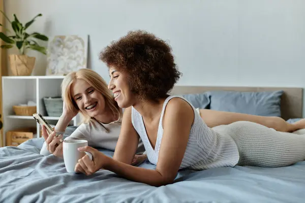 Ein lesbisches Paar entspannt sich im Schlafzimmer und lacht zusammen, während sie auf ihr Handy schauen.. — Stockfoto