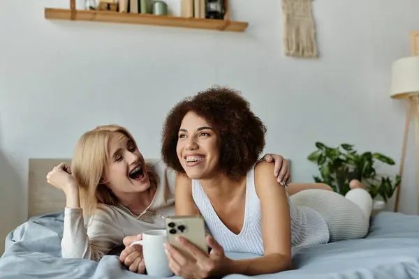 Una pareja de lesbianas se ríe juntos mientras están acostados en su cama, uno sosteniendo una taza, el otro un teléfono inteligente. - foto de stock