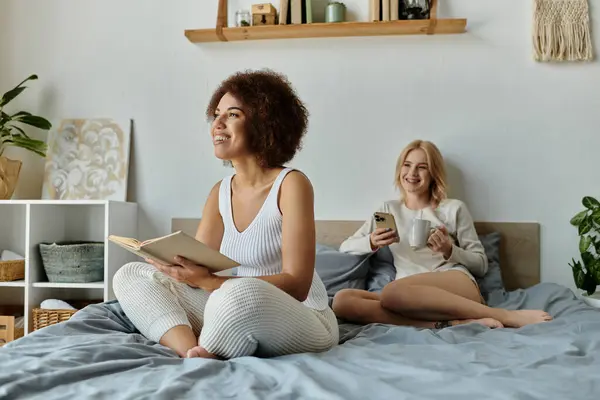 A couple relaxing in bed, one reading a book while the other scrolls through their phone. — Stock Photo
