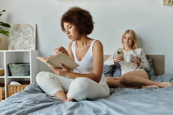 A multicultural lesbian couple relaxes at home, one engrossed in a book while the other scrolls on their phone. — Stock Photo