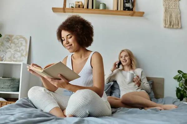 Un couple lesbienne multiculturel se détend à la maison, l'un lisant un livre et l'autre profitant d'un appel téléphonique. — Photo de stock