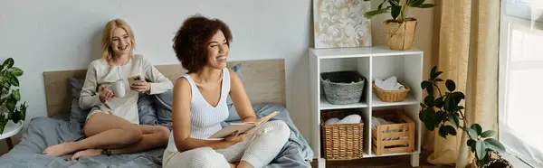 Two women in cozy clothes relax on a bed, sharing a moment of togetherness. One reads a book while the other sips from a mug — Stock Photo