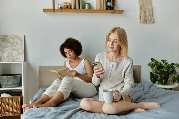 Due donne in abiti accoglienti rilassarsi su un letto, una lettura di un libro, mentre l'altra controlla il telefono e sorseggia una bevanda calda. — Foto stock