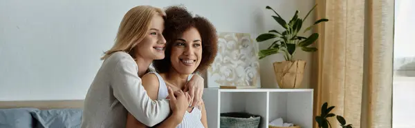 Due donne, una con i capelli biondi e l'altra con i capelli castani ricci, si abbracciano in un ambiente domestico caldo e invitante. — Foto stock