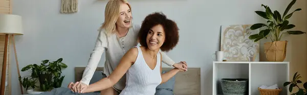 Two women, dressed in comfortable clothing, laugh and hold hands on a bed at home. — Stock Photo