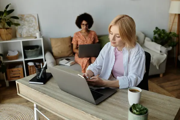 A lesbian couple works from home, one on a laptop and the other on her phone, while enjoying a cozy workspace with a cup of coffee. — Stock Photo