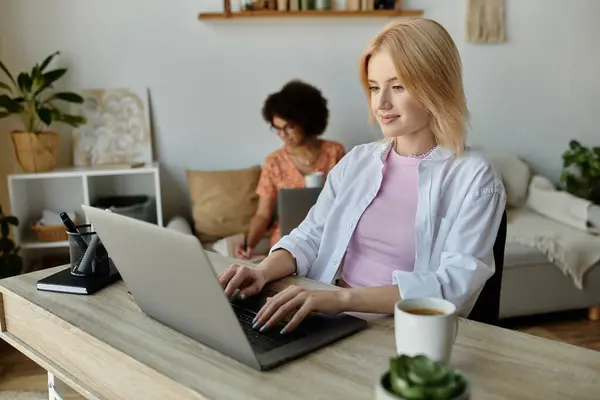 Ein lesbisches Paar arbeitet von zu Hause aus, eine Frau am Laptop, während die andere in der Nähe sitzt und sich auf ihre eigenen Aufgaben konzentriert.. — Stockfoto