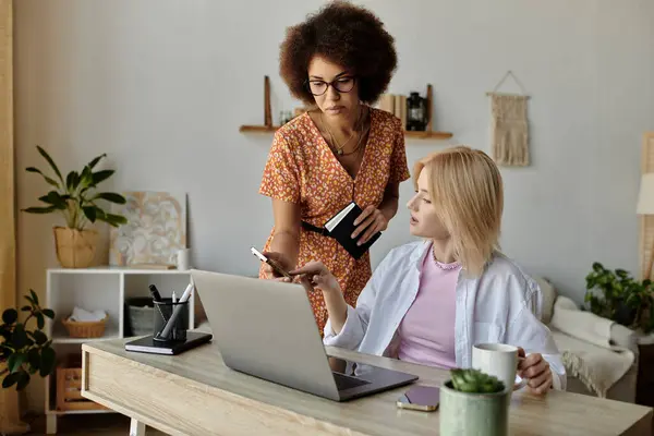 Duas mulheres trabalham juntas em uma mesa, uma apontando para uma tela de laptop, enquanto a outra segura uma caneca de café. — Fotografia de Stock