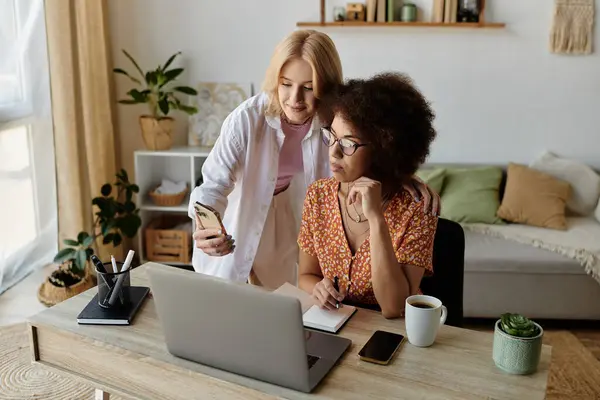 Two women, one holding a phone, look at a laptop screen while working from home. — Stock Photo