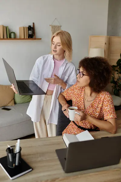 A lesbian couple works from home, one woman holds a laptop and points to the screen while the other sips coffee. — Stock Photo