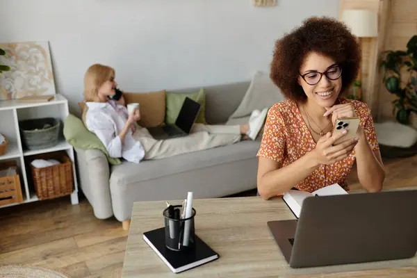 Eine Frau checkt ihr Handy, während ihr Partner auf der Couch im Hintergrund entspannt und ferngesteuert am Laptop arbeitet. — Stock Photo