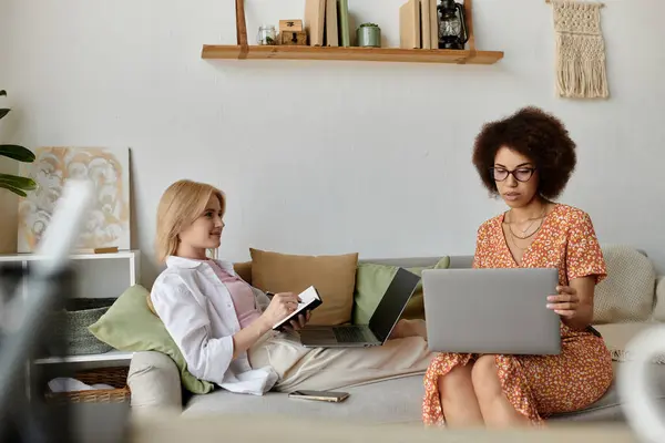 Two women, one blonde and one with curly brown hair, work on their laptops while sitting on a couch. — Stock Photo
