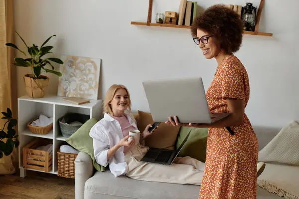 A lesbian couple working from home, one woman relaxing on the couch with a coffee, the other standing and holding a laptop. — Stock Photo